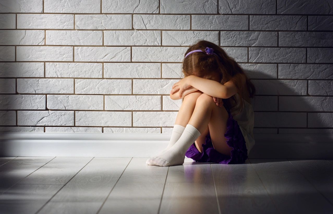 Young child huddled vulnerably against a white brick wall
